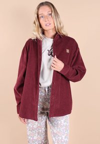 Chemise en velours côtelé bordeaux avec une coupe décontractée, ornée de boutons et d'un petit emblème. Assortie à un pantalon à motifs floraux.