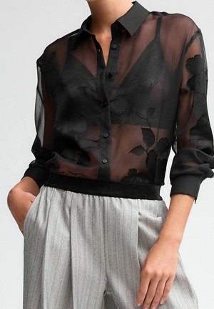 Femme portant un chemisier transparent noir à imprimé floral avec un soutien-gorge noir en dessous et un pantalon gris à rayures fines taille haute, mains dans les poches.