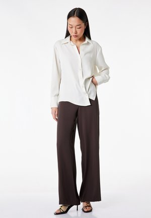 Vrouw staand, gekleed in een gebroken witte blouse met knopen, donkere wijde broekspijpen en zwarte sandalen met hoge hakken, hand gedeeltelijk in de broekzak.