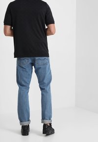 Černé polokošile s krátkým rukávem a texturovaným vzorem, kombinované s modrými denimovými džíny s ohrnutými manžetami a černými teniskami, z pohledu zezadu.