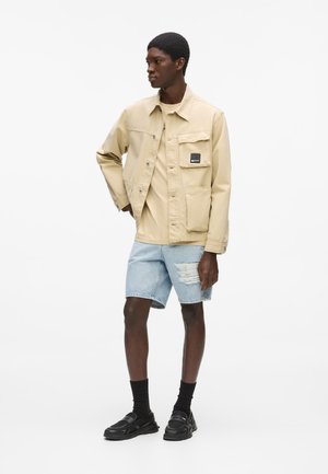 Beežist vabaaja jope nööbiga kinnituste ja rinnataskuga, kantud beeži t-särgi peal. Hele sinine kulunud teksapükstega lühike püksid ja mustad jalanõud.