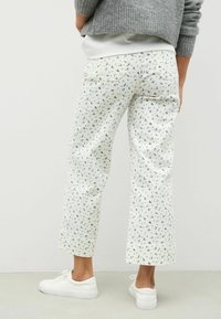 Pantaloni bianchi a cropped con un delicato motivo floreale blu, realizzati in tessuto leggero. L'outfit è abbinato a sneakers bianche e un maglione grigio.