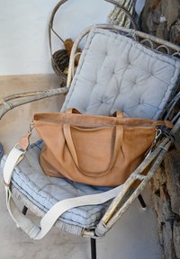 Brązowa skórzana torba tote z dwoma mocnymi uchwytami i odpinanym materiałowym paskiem na ramię, spoczywająca na wyściełanym krześle z szarym materiałowym pokryciem.