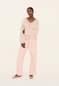 Strickpullover in Hellpink mit detailliertem offenen Muster und weiten Ärmeln, kombiniert mit lockeren, hellpinkfarbenen Hosen und beigen Ballerinas.
