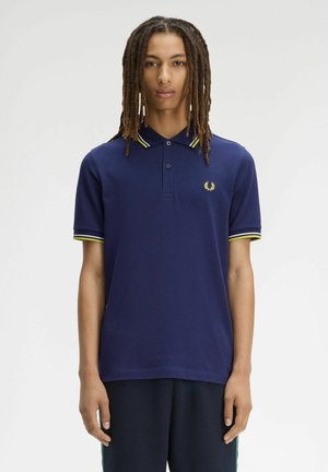 Joven con rastas largas que lleva una camiseta polo azul marino con cuello y puños de manga con rayas amarillas y blancas, de pie frente a un fondo liso.
