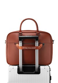 Bruine leren aktetas bevestigd aan een witte koffer met harde schaal en een telescopisch handvat, met de tekst "Victoria Hyde London" op de riem van de aktetas.