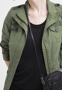 Giacca utilitaria verde oliva con dettagli a bottoni, chiusura con zip e tasche sul petto, indossata sopra una t-shirt nera a maniche corte. Piccola borsa a tracolla nera.