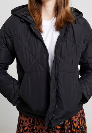 Frau trägt eine schwarze, gesteppte Kapuzenjacke über einem weißen Hemd und einem braunen Rock mit schwarzem und orangefarbenem Leopardenmuster.