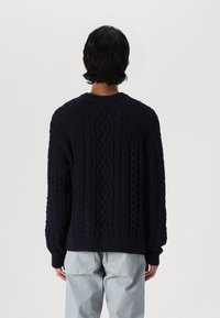 Marineblauer Kabelstrickpullover mit filigranen Mustern, Rundhalsausschnitt und langen Ärmeln. Kombiniert mit hellblauen Jeans, die ribbelige Bündchen zeigen.