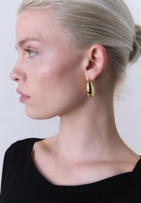 Goldene Tropfenohrringe, die an einem Modell präsentiert werden. Der Ohrring hat eine reflektierende Oberfläche und eine glatte, runde Form. Das Modell trägt ein schwarzes Oberteil.