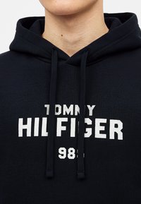 Човек, носещ черен суитшърт с качулка с отпечатано "Tommy Hilfiger 1985" в бяло на гърдите и черни връзки, които висят надолу.