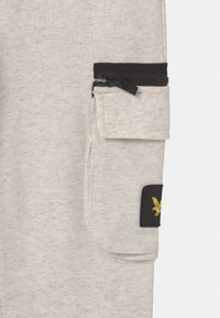 Lichtgrijze cargo-broeken met een zachte textuur, voorzien van een zwart met rits afgesloten zak en een geborduurd geel logopatch aan de zijkant.