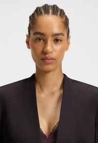 Sort blazer med en glat tekstur, der har en dyb V-udskæring over en lila top med blondekant. Håret er stylet i cornrows.