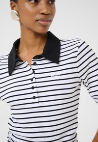 Svartvit randig poloshirt med svart krage, med knapplist och broderad detalj på vänster bröst.
