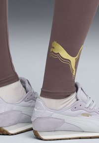 Beine in mauvefarbenen Leggings mit goldenem Puma-Logo und helllila Puma-Sneakern mit weißen Socken vor einem neutralen Hintergrund.