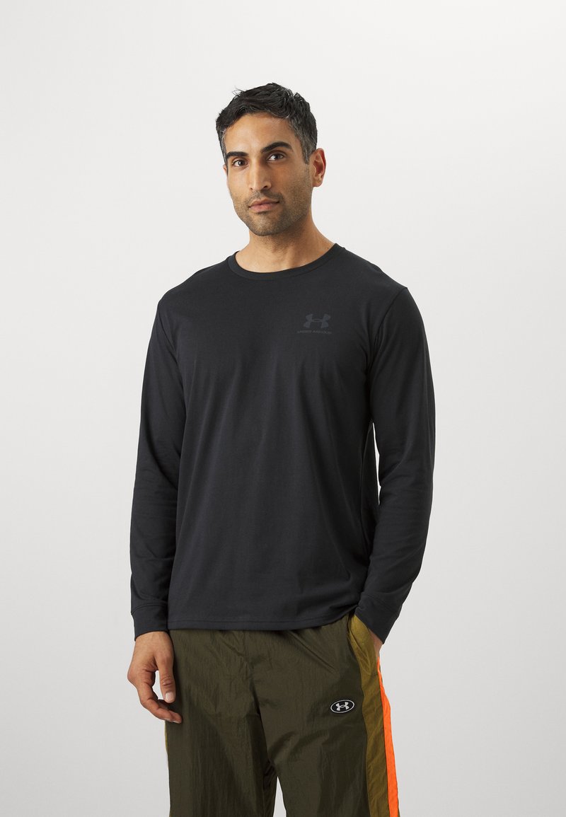 Schwarzes Langarmshirt mit rundem Halsausschnitt, versehen mit einem dezenten Logo. Getragen mit olivgrünen Hosen, die von orangen Streifen akzentuiert sind.