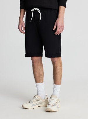 Mężczyzna ubrany w czarne spodenki na sznurki, białe skarpetki i beżowe trampki, stojący na gładkim jasnym tle.