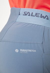 Gros plan sur la ceinture d'un pantalon de sport bleu avec le logo "SALEWA" et le logo "DURASTRETCH ACTIVE" sur le tissu près de la poche.