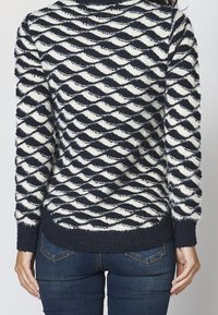 Svetr v námořnické modři a krémové barvě s vzorem vln, dlouhými rukávy a žebrovaným lemem, doplněný modrými džíny.