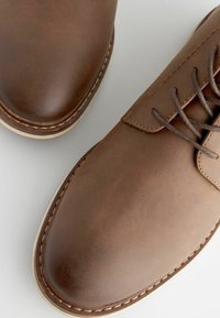 Zapatos de cuero marrón con una textura suave, puntera redondeada y suela de color claro en contraste. Presentan un laceado clásico y detalles de costura.