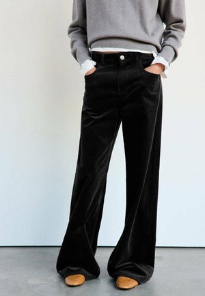 Persona que lleva pantalones negros de talle alto y pierna ancha, jersey gris con puños de camisa blancos y zapatos marrones, de pie sobre suelo gris.