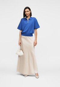 Μπλε μπλούζα πόλο με κοντά μανίκια, μπεζ μακριά φούστα και λευκή τσάντα. Το σύνολο έχει καθαρές γραμμές και χαλαρή εφαρμογή, με ομαλή υφή.