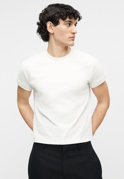 Νεαρός άνδρας με σγουρά μαλλιά που φοράει ένα απλό λευκό κοντομάνικο μπλουζάκι με στρογγυλή λαιμόκοψη και μαύρο παντελόνι, κοιτάζοντας προς τα δεξιά του.