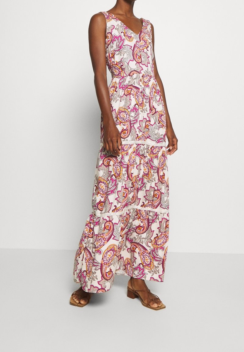 Femme portant une robe maxi blanche sans manches à col en V avec un motif cachemire rose, orange et gris, associée à des sandales marron à talons blocs ouvertes au bout.