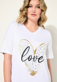 Bílý krátký rukáv tričko s výstřihem do V, zdobené zlatým a černým srdcem s nápisem "láska" a třpytivými detaily.