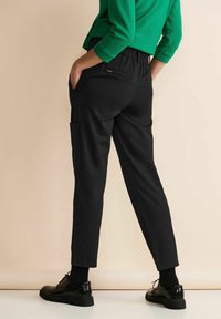 Person wearing black tapered pants with side pocket and black shoes, hands in pockets, standing against a beige background.