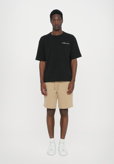 Tricou negru din bumbac cu o croială lejeră, asortat cu pantaloni scurți bej și adidași albi. Îmbrăcămintea are un design minimalist și proporții relaxate.