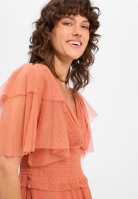 Perzikkleurige jurk met gelaagde tule mouwen, een smock-bodice en een v-halslijn. Getextureerde stof met subtiele rimpels bij de taille.