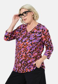 Monivärinen V-päänauha, jossa on kolmannes hihat, esittelee elävää abstraktia kuviota oranssissa, purppurassa ja mustassa. Valmistettu pehmeästä kankaasta.