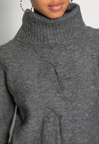 Femme portant un pull à col roulé en tricot gris et des boucles d'oreilles en argent, montrée de la mâchoire au torse moyen sur un fond uni.