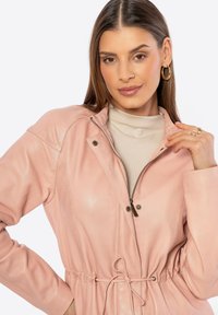Giacca in pelle rosa chiaro con chiusura a zip, vita con cordoncino, spalle strutturate e dettagli con bottoni a pressione. Texture liscia, vestibilità comoda.