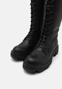 Černé vysoké boty z kůže s texturovaným povrchem, s předním šněrováním, zaoblenou špičkou a robustní podrážkou.