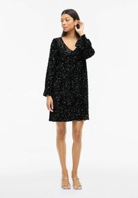 Juoda mini suknelė iš blizgančių karoliukų su ilgomis rankovėmis ir V formos iškirpte. Tekstūruota medžiaga su blizgančiais elementais sukuria atspindinčią paviršių.