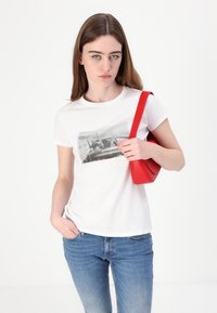 Bílý tričko s černým grafickým potiskem, nošené s modrými džíny. Modelka drží červenou, kulatou crossbody tašku s elegantním designem.