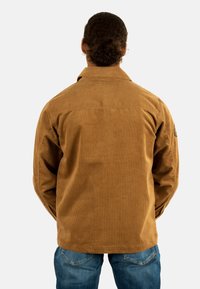 Chaqueta de pana marrón con estrías verticales, cuello abotonado y costuras detalladas. Lleva puesta sobre unos vaqueros de mezclilla azules, vista desde la parte posterior.