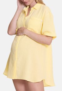 Geel hemdjurk gemaakt van textuurstof, voorzien van een knoopsluiting aan de voorkant, korte mouwen en een enkele borstzak. De zoom is iets gebogen.