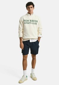 Crème sweatshirt met "NEW HAVEN SPORTS CLUB" in groene tekst, gecombineerd met marineblauwe shorts, een wit shirt eronder en crème sneakers met groene accenten.
