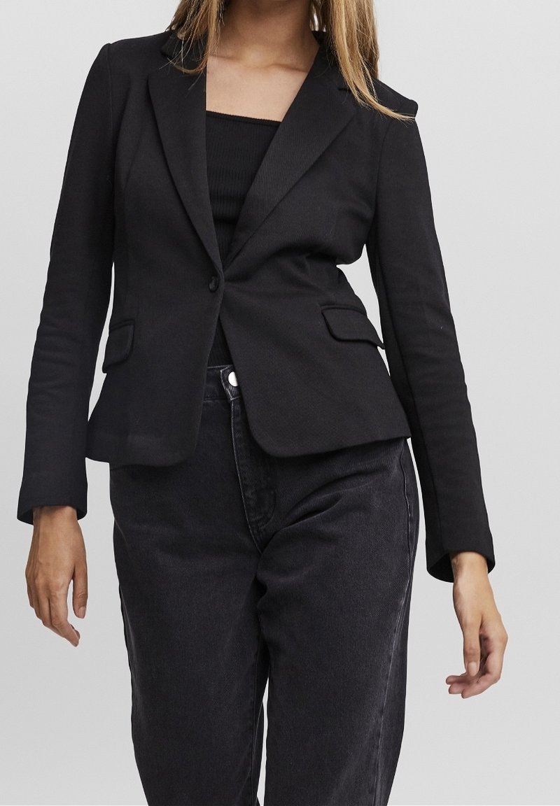 Zwarte op maat gemaakte blazer met een getailleerde snit, revers met inkeping, twee voorzakken en lange mouwen, gedragen over een zwart topje en donkere spijkerbroek.