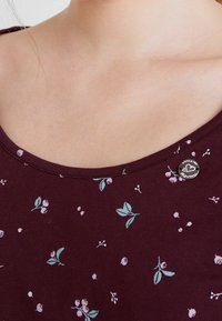 Bordeauxfarbener Oberteil mit einem runden Ausschnitt, verziert mit kleinen rosa Blumendrucken und grünen Blättern. Besitzt ein silbernes Etikett mit Schreibschrift-Details.