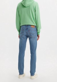 Personne portant un jean denim bleu clair et un sweat à capuche vert, debout de dos, avec des baskets blanches sur un fond blanc uni.