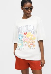 Άτομο που φοράει λευκό oversized t-shirt Adidas με πολύχρωμο γραφικό Care Bears και κόκκινα σορτς Adidas, αξεσουάρ με μπεζ γυαλιά ηλίου.