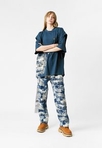 Joven de pie con los brazos cruzados, vistiendo una camiseta suelta de color azul marino, pantalones de patrón azul y blanco, y zapatos de plataforma color tostado, contra un fondo blanco.