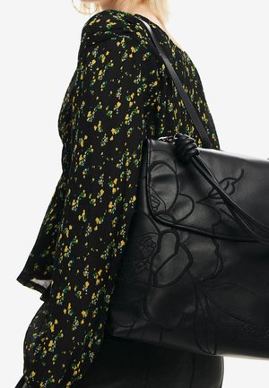 Musta nahkainen käsilaukku, jossa on kohokuvioitu kukkakuvio, kiinnitetty kiertytyneellä hihnalla, käytetty yhdessä mustan kukkakuvioisen paidan kanssa, jossa on keltaisia yksityiskohtia.