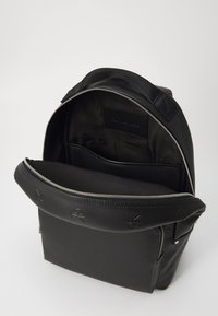 Zaino nero realizzato in materiale sintetico liscio. Presenta un'apertura con zip, tasche interne e dettagli del logo in rilievo all'esterno.