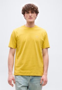 Νέος άνδρας με σγουρά μαλλιά που φοράει κίτρινο T-shirt BOSS και ανοιχτόχρωμο τζιν, στέκεται μπροστά σε απλό ανοιχτόχρωμο φόντο.