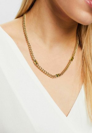 Eine goldfarbene Kettenhalskette mit ineinandergreifenden Gliedern und rechteckigen Akzenten, kombiniert mit einem weißen Oberteil und langen, glatten Haaren.